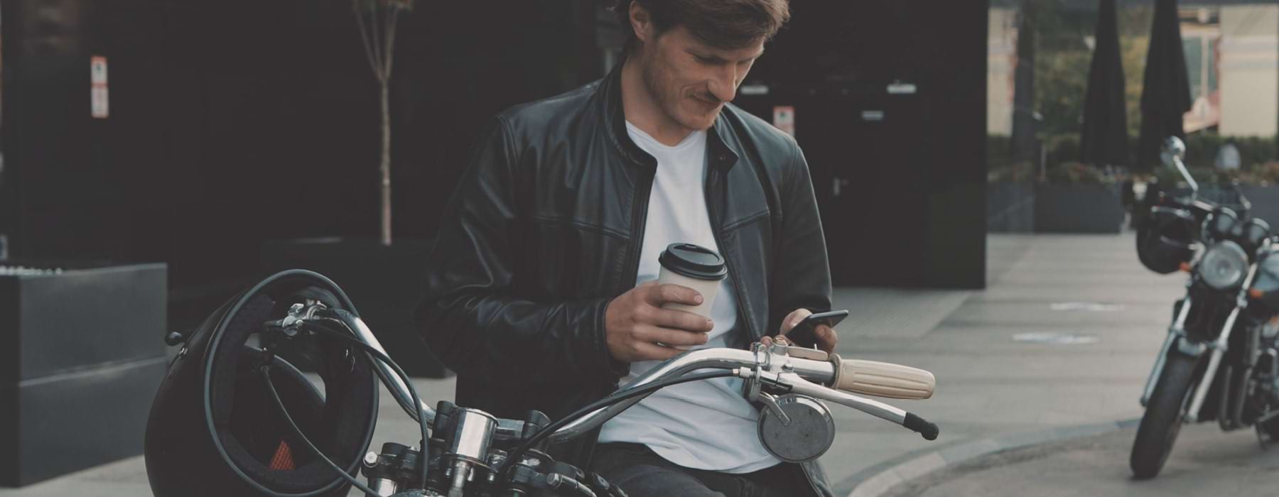man sits on parked motorcycle and texts with cup of coffee in hand